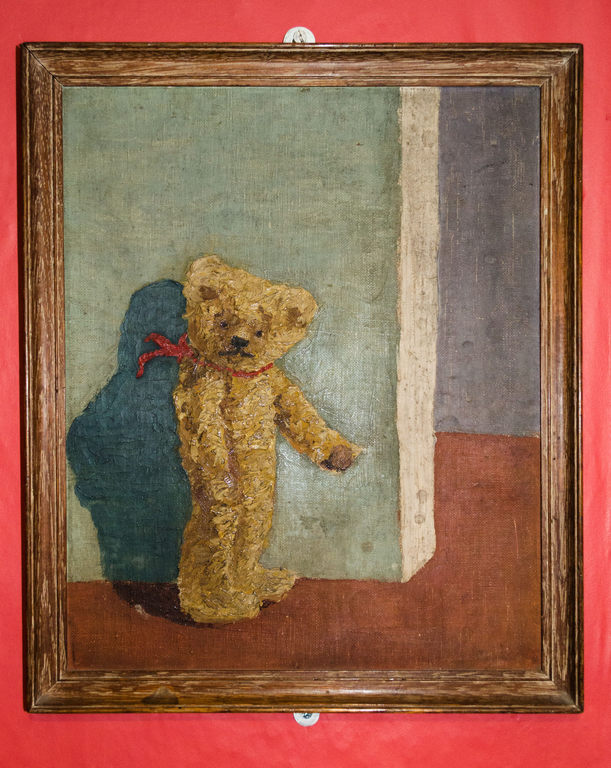 A droopy-eyed teddy bear that may have been painted with cigarette butts stands before a bare wall, limply gesturing around a corner which appears to lead only to more emptiness.