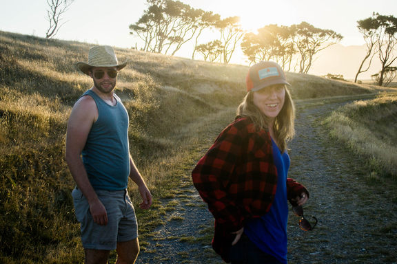 Sam and Britt turning back and smiling on a dusty road at sunset. Some neat trees behind.