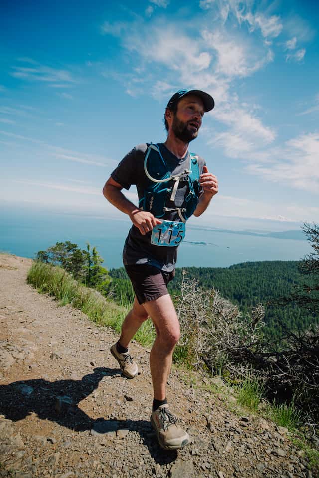 A head-to-toe portrait of me, in three-quarters view, running on a trail on a bright sunny day. There’s a beautiful view of the sea and islands behind me. I’ve got a bright, wide-eyed expression, like the tail end of a smile, and am looking straight ahead.