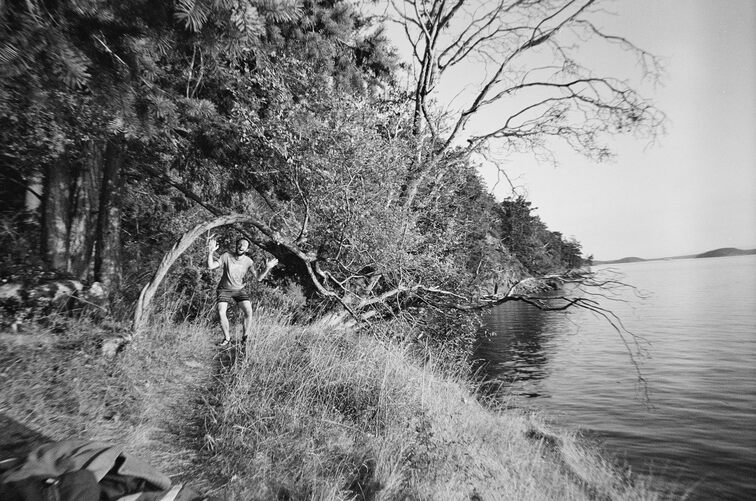 A man (me), crouching under a semi-circular twisted branch, on a cliff above the sea