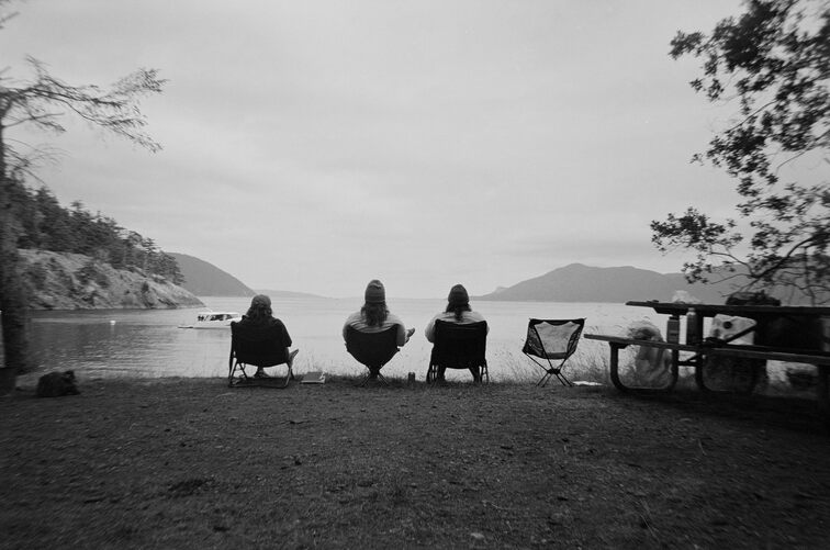 Four camp chairs on a grassy knoll, facing a cover, and the sea, and some islands. Three of them are occupied, with people watching the water. One is empty.