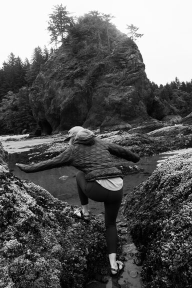 Britt, photographed from behind, climbing over barnacled rocks on a beach with a big haystack in the background. She's pretty close to the camera, and about to prop herself up with her left hand, which is fully extended.