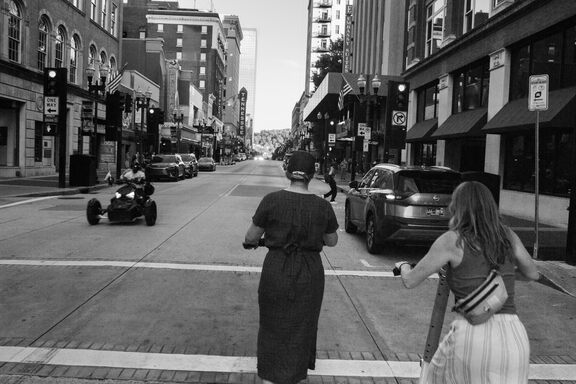 Two women photographed from behind, scooting on scooters down a main street in a downtown area. In the oncoming lane is a weird-looking three wheeled vehicle.