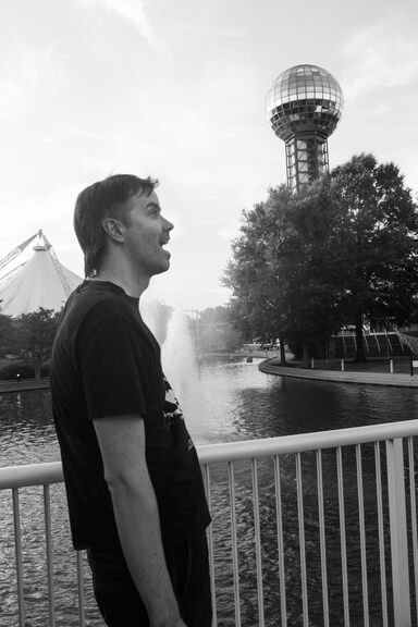 A man (Justin) stands agape in the foreground, pantomiming amazement, while a very modestly-sized 80s-looking piece of decorative architecture (the Sunsphere) sits behind him in the middle distance