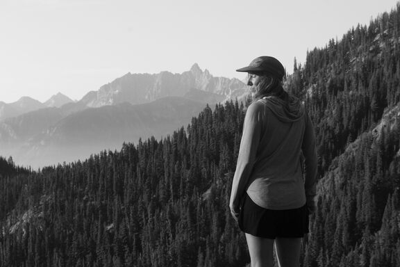 Britt, with her body facing away but with her face turned back towards the camera - eyes closed, smiling - in front of a jagged mountain range in the distance