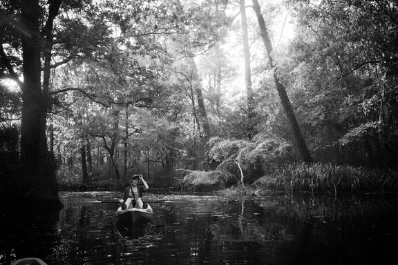 Britt, in a kayak, on a glassy swamp, tipping her baseball cap, beneath a canopy of large trees.