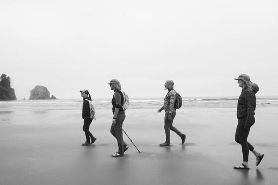 Four people walking on a beach. They're in cold-weather clothing; it's misty, there are a couple of large rock formations sticking out of the water in the distance.